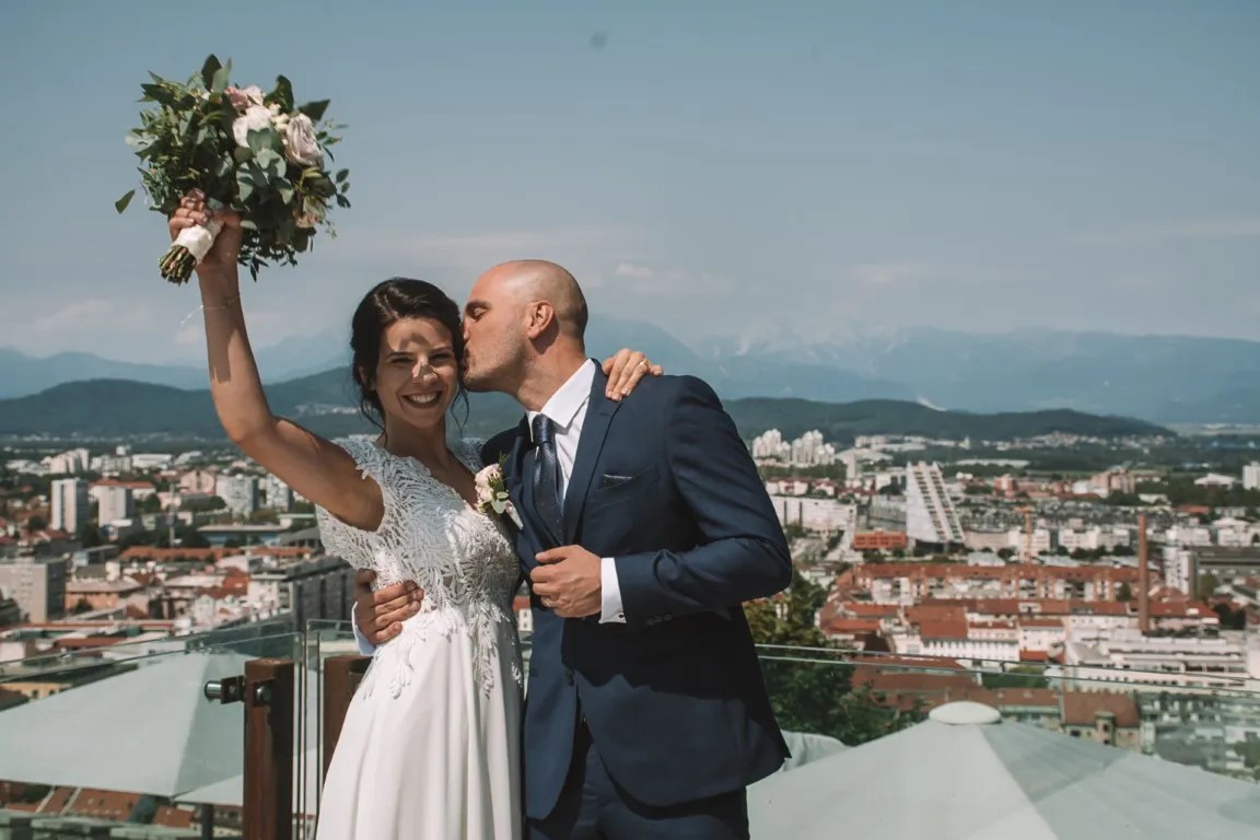Mladoporočenca na Ljubljanskem gradu v Ljubljani; intimen trenutek ob grajskem obzidju v dokumentarnem poročnem slogu s pogledom na mesto.